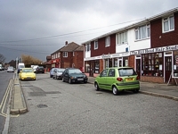 An image of a row of shops on the right side with cars parked in front in a parking area.