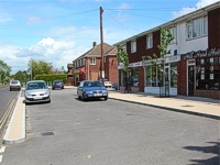 An image of the same row of shops and parking area showing new road surfaces and newly planted trees.